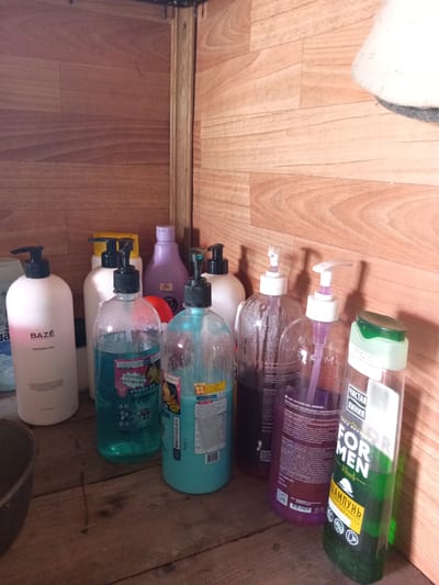 A collection of personal care product bottles, primarily shampoos and shower gels, are arranged on a wooden surface. The bottles are of various shapes, sizes, and colors, with several featuring pump dispensers. The background consists of wooden paneling. The scene appears to be an indoor storage area, possibly a bathroom or utility space, located in Monastirische, Russia.
