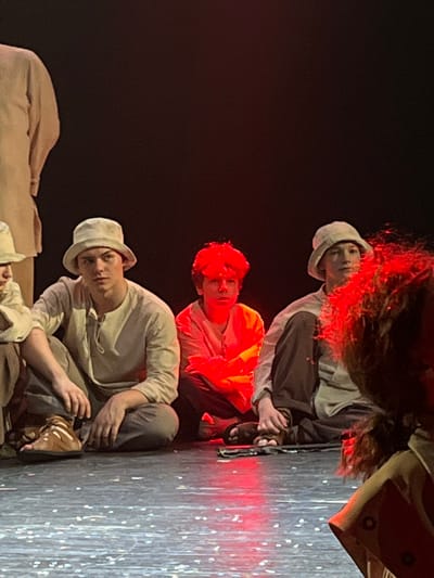 The image depicts a theatrical scene on a stage in Novoshakhtinsk, Russia, featuring five individuals. In the mid-ground, three young males are seated on the dark stage floor, facing generally forward. The individual on the left, a young adult with a light-colored bucket hat and beige tunic and trousers, sits with hands resting on his knees and wears brown sandals. He looks slightly to his left. The central individual, a younger male, is intensely illuminated by red light, making his face and reddish hair prominently bright red. He wears a light-colored tunic and dark trousers and sits with his legs crossed. The individual on the right, another younger male with a light-colored bucket hat and a light tunic with dark trousers, sits with his legs crossed and wears dark sandals. He looks slightly to his right. A partial view of a standing person in a light-colored tunic is visible on the far left, behind the seated individuals. In the immediate foreground on the right, the back of a person's head with voluminous dark, curly hair, also illuminated by red light, partially obscures the rightmost seated individual. The stage floor is dark and reflective, showing scattered light specks and red reflections from the central lighting. The background is uniformly dark.