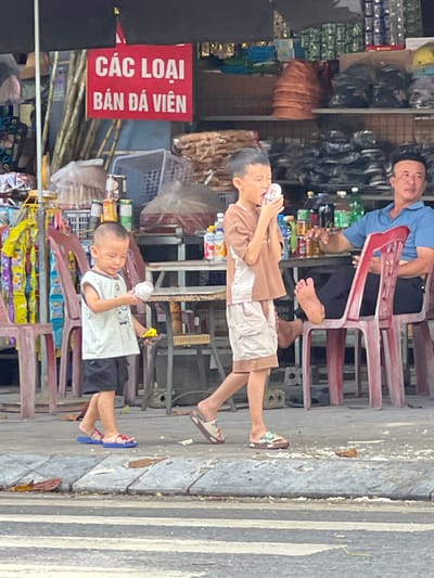 The image captures a street scene in Tân Quang Commune, Vietnam, depicting two young boys walking on a sidewalk. The older boy, wearing a brown t-shirt and light-colored shorts, is in the foreground, holding a white object to his mouth. The younger boy, in a white sleeveless shirt and black shorts, is to his left, holding a white cylindrical object and a yellow toy. In the background, a man in a blue shirt sits on a red chair. A stall with various items and shelves is visible behind the children and the man, featuring goods such as baskets, bags, and bottled drinks. A red sign with white Vietnamese text is prominently displayed above the stall. The ground is paved with concrete and a pedestrian crossing is visible in the lower portion of the frame. Fallen leaves are scattered on the sidewalk.