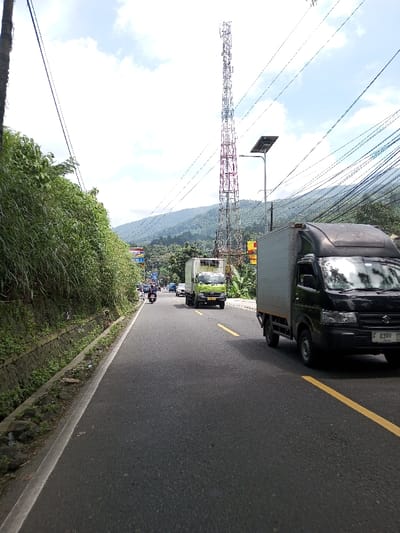 The image depicts a rural or semi-urban road scene in Boommlog, Bhutan, under daylight with a partly cloudy sky. The asphalt road features a solid white line on its left edge and broken yellow lane markers, indicating at least two lanes of traffic. Along the immediate left edge of the road is a concrete gutter, followed by a steep embankment densely covered in tall green vegetation. A utility pole is partially visible on the far left. Overhead, numerous power lines and communication cables traverse the frame.
In the foreground, a black box truck with a light grey cargo box is positioned in the right lane, oriented away from the viewer. A partial license plate "F 8399 N" is visible on its front. Further down the road, a green box truck, possibly with an open-top or caged rear section, is in the opposing lane, approaching the camera. Behind the green truck, a motorcyclist on a dark motorcycle is also visible, moving towards the camera. Additional distant vehicles are discernible further along the road.
In the mid-ground, to the right of the road, a tall, multi-segment lattice communications tower with red, white, and blue sections stands prominently. Adjacent to the tower is a street light pole topped with a solar panel. Beyond the immediate road and tower, the landscape features rolling hills and mountains covered with dense green foliage, receding into the distance.