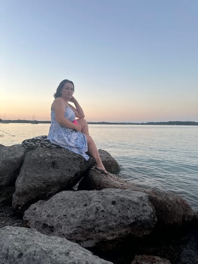 A woman is sitting on rocks by the water at sunset. She is wearing a blue and white dress and has her hand on her chin. The sky is a light blue and pink, and the water is calm. There are sailboats in the background. The location is the shore of the Chiemsee in Prien am Chiemsee, Germany.