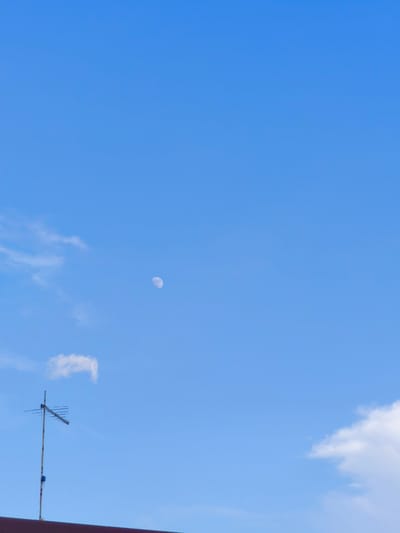 The image displays a clear blue sky with a few wispy clouds. In the upper left quadrant, a crescent moon is visible. Towards the bottom left, an old-fashioned television antenna stands tall on top of a building, its metallic structure silhouetted against the sky. A small, fluffy white cloud drifts above the antenna. The scene suggests a daytime setting, likely mid-morning or early afternoon, given the bright, clear sky. The location is identified as Chiugiana, Italy. There is no text visible in the image. The overall mood is serene and peaceful, with the vastness of the sky dominating the frame. The antenna hints at a more traditional, possibly older, setting in a residential or rural area. The lighting indicates fair weather.