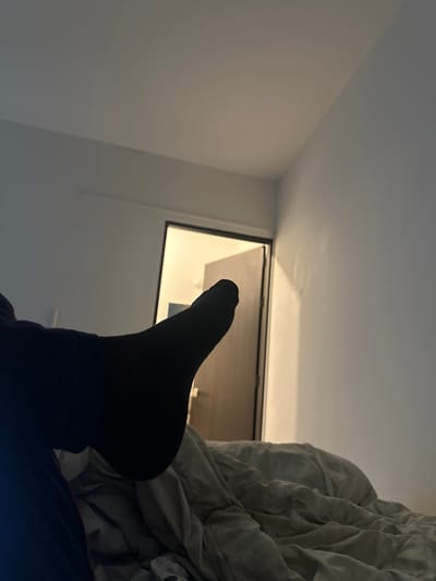 A low-angle perspective features a foot clad in a black sock pointing towards an open window. The window frame is dark, with bright light streaming through the opening. A rumpled white blanket partially obscures the view on the right side. The room consists of white walls.