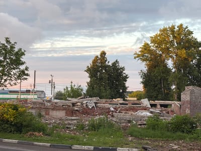 The image depicts the ruins of a building in Balabanovo, Russia, under a cloudy sky with hints of pink and orange from a sunset. The foreground shows a curb and a grassy area. The midground is dominated by the rubble of a destroyed brick building, with concrete slabs, broken bricks, and twisted metal scattered amongst overgrown vegetation. In the background, several trees are visible, with lush green foliage. Behind the trees, there is a long, low building with a white and green facade, and further in the distance, more trees line the horizon. Electrical poles and wires are also present in the background.
