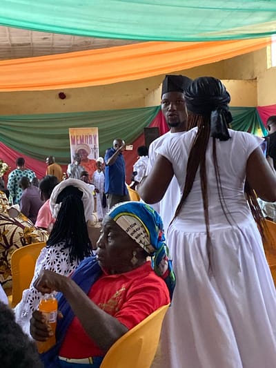 The image depicts an indoor gathering, likely a memorial service or celebration, in Kuru, Nigeria. The space is decorated with draped fabrics in green and orange, creating a festive but somber atmosphere. In the foreground, a woman in a red shirt and blue headscarf is seated, holding a bottle of orange liquid. She appears to be looking to her right. Behind her, a woman in a white dress with long braids is partially visible, facing away from the camera. A man in a black cap and white shirt stands next to her, looking to his left. In the background, a banner with the word "MEMORY" and a picture of a woman is displayed. Several other individuals are present in the scene, some seated and others standing. The lighting is somewhat dim, suggesting an indoor event. The overall impression is one of a community coming together for an occasion.