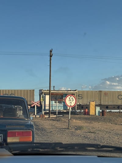 In Töretam, Kazakhstan, at a railroad crossing, a dark blue vehicle is stopped, seen from behind. The vehicle's taillights are visible, one red and one orange. Beyond the car, a railway crossing sign with red and white stripes is present, along with a circular traffic sign indicating a height limit of 3.5 meters. A small building, possibly a guardhouse or station, is situated to the right of the crossing sign. In the background, long, ribbed metal containers are stacked and arranged horizontally, suggesting a freight or cargo area. A wooden utility pole with wires stretches across the scene, and the sky above is clear blue with some scattered clouds. The ground is a mix of dirt and gravel.