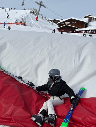 The image captures a snowy mountain ski resort scene, specifically identified by the "PAS DU LAC" sign, indicating the Pas du Lac area within Méribel, part of Les Allues, France. In the foreground, an individual is seated on a red inflatable safety barrier or cushion. This person is wearing a grey ski helmet with goggles, a black insulated jacket, white ski pants, and black ski boots. They are holding a pair of blue and green skis, one on each side, with their gloved hands resting on the ski bindings. A large white inflatable structure forms a backdrop directly behind the seated individual.
The midground features a wide, snow-covered ski slope where multiple skiers are visible, actively skiing or standing. A large metal ski lift pylon with a red base is positioned on the slope to the left. A red safety fence or netting runs across the lower part of the slope. To the right, a prominent multi-story wooden chalet-style building, marked with the sign "PAS DU LAC," stands with snow-covered roofs, suggesting a restaurant or lodge.
In the background, more snow-covered slopes rise, dotted with evergreen trees. A chairlift system with moving chairs and additional skiers is visible further up the mountain. The sky is uniformly overcast. The scene depicts a busy winter sports environment.
