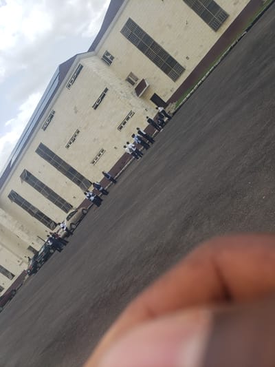 The image depicts a group of individuals, likely students or staff, in uniform standing in a line outside a modern, beige-colored building with a dark-tiled roof. The building features multiple rows of windows, some with metal grating. The ground is paved with asphalt, and there are several cars parked in a line. The scene appears to be an outdoor area in front of an institution, possibly a school or office complex. The sky is overcast with clouds. The angle of the photo is tilted, suggesting it was taken from a low vantage point or with the camera held at an odd angle. The location is identified as Kachia, Nigeria.