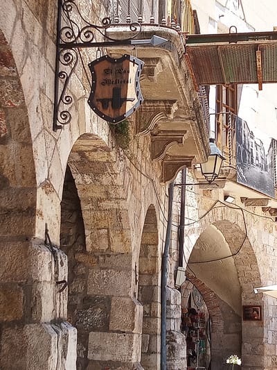 The image captures a section of a street in Besalú, Spain, characterized by its historical stone architecture. In the foreground, a weathered stone wall with multiple arches dominates the scene. Ornate wrought iron brackets support a shield-shaped sign hanging above the arches. The sign appears to be made of wood with metal accents and displays text and a graphic, possibly advertising a medieval-themed establishment. A vintage-style street lamp is mounted on a balcony above the arches. The background reveals more of the building's facade, including balconies, windows, and a mural depicting a cityscape. Wires are strung across the scene, typical of older urban environments. The overall impression is one of an old European town with a focus on historical preservation.