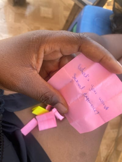 A close-up image taken in Tudun Nupawa, Nigeria, shows a dark-skinned hand holding a creased, pink square piece of paper. Blue handwritten text on the paper reads: "What's your worst sexual experience?". The hand's thumb and index finger are visible on the left side of the paper. Below the hand, on a dark fabric, are several small, folded pieces of paper; most are pink, and one is yellow. The dark fabric appears to be clothing, with a visible braid suggesting it is a person's upper leg. The background shows a light-colored, possibly tiled floor and a partially visible blue plastic object, likely a chair or stool. The scene is brightly lit.