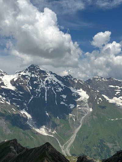 This is a vertical, eye-level, medium shot of the Großglockner mountain in Fusch an der Großglocknerstraße, Austria, on a partly cloudy day. The mountain's summit is covered in snow and ice, with dark rocky slopes descending into green alpine meadows dotted with trees. A glacier flows down one side of the mountain, feeding a dry riverbed. The sky is a vibrant blue, with dramatic white clouds casting shadows on the mountainside. The foreground features the jagged peaks of a lower mountain range covered in green grass and dark rocks. There are no people or animals visible in the image. The overall scene depicts a majestic and rugged alpine landscape.