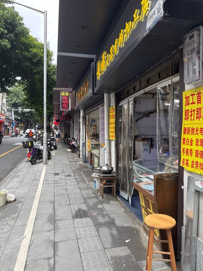 The image depicts a street scene in the Liwan District of China. On the right side of the image is a storefront with a dark facade and bright yellow signage. The signage appears to be in Chinese characters and is illuminated. Below the signage, a transparent curtain hangs across the entrance to the shop, and a glass display case is visible inside. A wooden stool stands on the sidewalk in front of the shop. To the left of the storefront, a sidewalk is paved with gray tiles. Several electric scooters are parked along the sidewalk. Further down the street, trees line the road, and traffic signals are visible in the distance. The sky is overcast. A person is seated on a small stool on the sidewalk in front of the shop. The scene appears to be a commercial area with shops and pedestrian traffic.