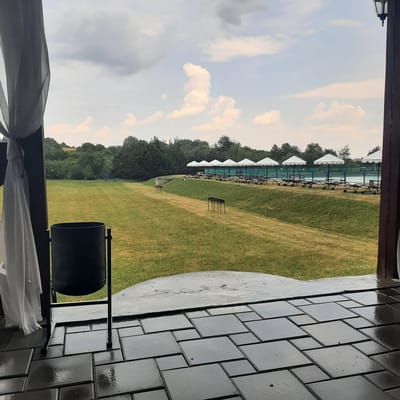The image depicts an outdoor scene at a facility in Dubrivka, Ukraine. In the foreground, a dark, wet patio is laid with rectangular and square dark gray tiles. A black trash receptacle stands on the patio. A white curtain hangs from the left side of the frame. Beyond the patio, a lush green lawn slopes upwards. Several metal barbecue grills are placed on the lawn. Further back, a row of white umbrellas shelters lounge chairs. A blue fence borders a body of water behind the umbrellas. A dense line of trees forms the background, with a hint of hills and sky visible above. The weather appears overcast, with visible rain drops in the air.