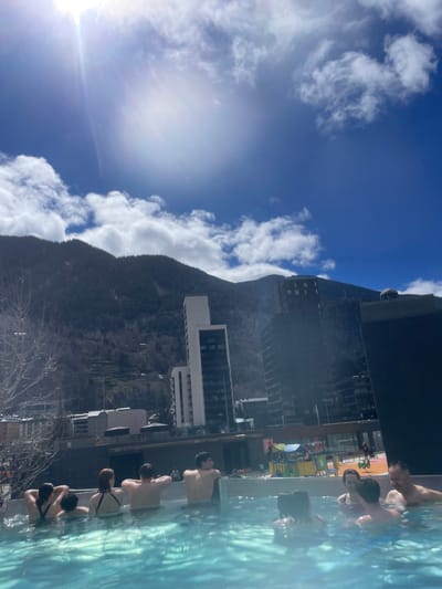 The image, taken on a sunny day with a blue sky dotted with puffy white clouds, shows a scene in Les Escaldes, Andorra, capturing the city's unique setting amidst mountains and modern architecture. A steep, dark mountain backdrop dominates the left side of the frame, contrasting sharply with the bright sky. Modern buildings, including tall apartment blocks and hotels, cascade down the mountainside. On the right side, several people are enjoying an outdoor pool. They are interacting, bathing, and relaxing while enjoying the view of the surrounding landscape. The pool water is a light blue and appears refreshing.