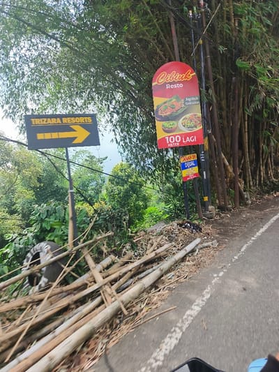 The image depicts a roadside scene in Sukasari, Indonesia, located at coordinates -6.837 latitude and 107.598 longitude. A black sign with white lettering reading "TRIZARA RESORTS" and a yellow arrow pointing right is mounted on a metal pole on the left side of the image. A second pole on the right side of the image displays an advertisement for a restaurant called "Cibluk Resto," with a picture of food and the phrase "100 LAGI METER." The pole also has a smaller sign beneath it. The foreground is covered in broken bamboo branches. In the background, there is a paved road with a white stripe running down the center. The road is flanked by dense foliage on both sides.