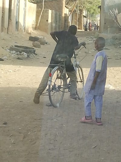 In a dusty street setting, a person in dark clothing leans over a bicycle with a flat tire, while another child, dressed in light-colored clothing and pink sandals, stands to the side looking directly at the camera. The background shows buildings and other people in the distance. A discarded tire sits near a pile of debris.