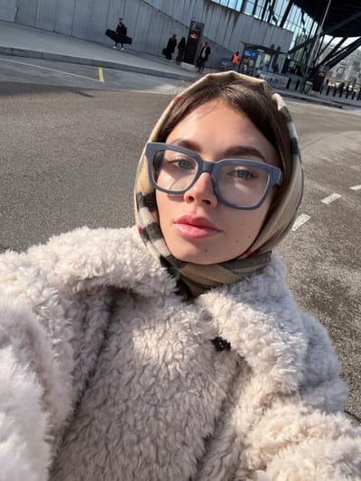 A young adult female is depicted in a close-up, upward-angled selfie, standing outdoors at Lyon-Saint Exupéry Airport in Colombier-Saugnieu, France. She wears grey-framed rectangular glasses, a light beige shaggy coat, and a beige plaid scarf wrapped around her head and neck. Her hair is dark, and her lips appear lightly colored. The background features an asphalt surface with white and yellow painted lines. In the mid-ground, a concrete wall and a vertical dark grey sign displaying the number "2" are visible. Further in the background, multiple individuals are present, one carrying a large, dark rectangular case. A structure resembling an information kiosk with a blue banner is also visible, along with multiple luggage trolleys. Behind these elements, a modern airport terminal building with extensive glass and steel facades is present. The scene is illuminated by bright daylight.