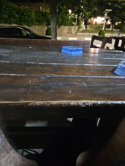 The image depicts an outdoor setting, likely a cafe or restaurant patio, at night. A dark wooden table occupies the foreground, showing visible wear and scratches. On the table, a blue ashtray is visible on the left, and a portion of another blue object, possibly another ashtray or part of a seating area, is seen on the right. In the background, trees with foliage are illuminated by ambient light, suggesting streetlights or building lights. A dark-colored sedan is parked on the left side of the frame, partially obscured by the table. The overall scene is dimly lit, with a focus on the textured surface of the table. The mention of Biaga, Bulgaria, is external context and not visually verifiable from the image itself.
