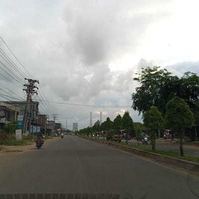 This image depicts an overcast street scene in Pantegajah, Indonesia, captured from a moving vehicle. The sky occupies the upper half, dominated by thick, gray clouds with brighter areas suggesting diffuse sunlight on the right.

In the foreground, the asphalt road surface is visible, dark gray and appearing slightly damp or textured. There are no clear lane markings.

On the left side of the road, a dense network of black electrical or communication wires spans the upper left quadrant, attached to a prominent utility pole with multiple cross-arms and insulators. Below the wires, a row of two-story buildings lines the street, some appearing to be shops or residences, constructed from concrete or brick. A sign displaying "PLN" (Perusahaan Listrik Negara, Indonesian state electricity company) is visible on one of these buildings. A small white informational sign stands on the unpaved verge in front of the buildings. A motorcyclist in dark attire is visible on the road, heading away from the camera, roughly in the center-left. Other motorcycles are visible further down the road on the left side.

On the right side of the road, a raised concrete curb separates the asphalt from a strip of green grass and a continuous row of neatly trimmed, conical evergreen trees. Behind these smaller trees, larger, irregularly shaped trees with dense green foliage are visible. Further down the road, behind the trees, various vehicles including a light-colored truck and a three-wheeled vehicle with an illuminated frame can be discerned.

In the mid-distance, the most prominent features are two tall, slender white mosque minarets topped with golden domes, positioned centrally in the frame. A darker dome structure of the mosque is situated between and slightly in front of the minarets. To the left of the mosque, a tall, slender telecommunication tower or mast is visible on the horizon, alongside a large, rectangular billboard structure.

The overall environment suggests a semi-urban setting, characterized by mixed commercial and residential structures, overhead utility lines, and common road traffic, predominantly motorcycles. The cloudy sky casts a soft, uniform light over the entire scene.