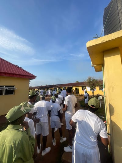 In Emure-Ekiti, Nigeria, a group of young people, predominantly male, are lined up outside a yellow building under a clear blue sky with scattered clouds. Many are wearing white t-shirts and shorts, some with green caps. The building has a red-tiled roof and open windows. A large black water tank is visible on the roof of another yellow structure. In the foreground, a person in a green uniform jacket and cap is facing away from the camera. The overall scene suggests an outdoor gathering or activity in an institutional setting.