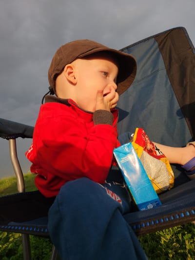 A young child is sitting in a blue and black camping chair. They are wearing a red jacket and a brown hooded cap. The child is looking to the left and has their fingers near their mouth, as if sucking on them. Beside the child, there is a package of snacks, one is yellow and red and the other is blue. The child's legs are bent and they are wearing blue pants. The scene is outdoors, with green foliage visible in the background and a cloudy sky above. The lighting suggests it might be late afternoon or early evening. The image is oriented vertically.