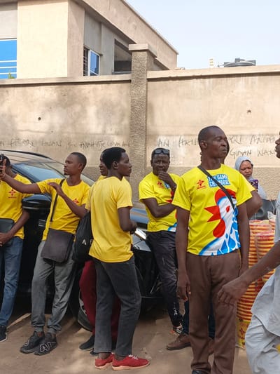 This outdoor scene in Kawaji, Nigeria, captures a group of people, predominantly men, gathered during the daytime under clear, sunny skies. Most of the men wear bright yellow t-shirts, indicating their association with the "MAGGI" brand, likely for a promotional event. Several shirts display the "MAGGI" logo, while one man in the foreground wears a distinctive t-shirt featuring "MAGGI GAME CHANGER!" and a vibrant rooster graphic in red, white, and blue.
The group is situated next to a black car, with a concrete building wall forming the background. One man on the far left is holding a smartphone, possibly capturing a photo or video. Another woman in a grey head covering is visible in the background. On the right, stacks of yellow plates or bowls, some bearing the "MAGGI" brand, are visible.
The concrete wall behind the individuals is marked with graffiti, including phrases like "A FITSARI ANARI BAN ALLAH ATE CICAN WAIK" and "AN HAN" with the number "04 PE 2383506". The architecture and casual attire are consistent with the Nigerian setting. The overall mood appears calm, with individuals either observing or engaging in conversation.