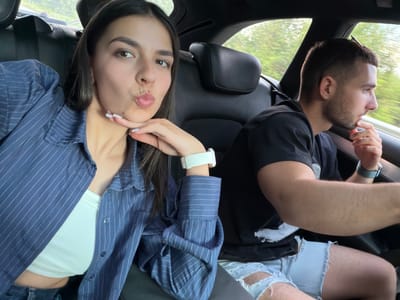 The image, possibly taken from the interior of a moving vehicle in Брусовeте, Bulgaria, captures two individuals. The person on the left appears to be a man, with short hair and a beard, wearing sunglasses and a black t-shirt. A wrist watch is visible on his left wrist. His lower half is covered by a pair of light blue ripped jeans. To the right, a woman with dark hair and fair skin is positioned. She is wearing a white top underneath a blue pinstriped button-down shirt. She has a white smartwatch on her left wrist. Her expression is a "duck-face" pout. The dark interior of the vehicle forms the background, and glimpses of greenery are visible through the window.