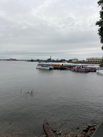 The image captures a waterfront scene in Kronstadt, Russia, under an overcast sky. In the foreground, the murky water laps against a rocky shore, with some reeds growing in the shallow areas. A rusted piece of metal debris is partially submerged near the bank.

Several boats are docked along a pier extending into the water. The most prominent is a white passenger boat with a blue and red stripe, featuring Russian text and an orange life preserver. Next to it, a longer vessel with an orange roof and a white cabin is moored. Further along the pier, a smaller, narrower boat with a blue and red stripe is also docked, and a metal gangway connects it to the shore.

In the middle ground, the water stretches towards a distant shoreline. A cluster of submarines is visible, suggesting a naval presence. Further in the distance, a lighthouse stands near some buildings, possibly part of a historical complex or naval facility. On the right side of the image, a large, yellow building with a long colonnade is situated along the waterfront, with a portion of a dark green tree visible in the upper right corner. The overall atmosphere is somber, indicative of the cloudy weather.