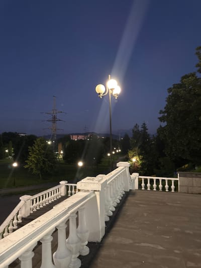 The image depicts a nighttime scene in Arkabash 1-Y, Russia. The foreground features a white decorative railing, characteristic of a park or public walkway. Beyond the railing, a path curves through a park area, illuminated by several lampposts that cast pools of light. In the mid-ground, a large transmission tower stands tall, its structure outlined against the dark blue twilight sky. Trees dot the landscape, their leaves appearing dark and dense in the low light. In the distance, faint lights suggest buildings or a settlement nestled among rolling hills or mountains. The sky is a deep twilight blue, indicating that it is either early evening or late evening. The overall mood is serene and quiet, with the artificial lights providing a warm contrast to the fading daylight. There is no visible text in the image.
