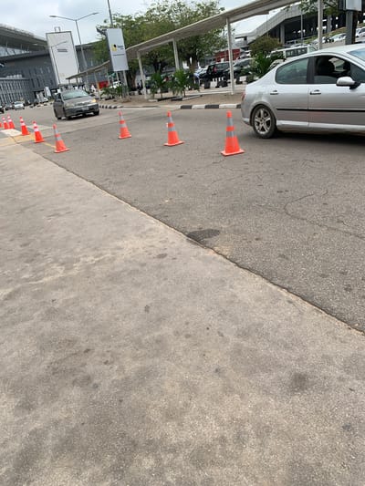 The image depicts a bright, sunny daytime scene on a roadside in Gwaska, Nigeria. A light-colored four-door sedan, possibly white or silver, is parked or slowly moving on the left side of the frame, positioned on an asphalt road. A distinct line of orange and white traffic cones stretches diagonally from the foreground to the mid-ground, serving as a barrier or demarcation along the edge of the asphalt. These cones separate the main roadway from a lighter, grey concrete surface, likely a sidewalk or shoulder, which occupies the right half of the image.
Further down the road, beyond the first few cones, a dark-colored sedan with noticeable damage to its front bumper is visible. In the background, tall modern buildings with glass windows and some scaffolding can be seen under a clear sky. Lush green trees and various plants line the road and buildings. Multiple streetlights and poles are scattered throughout the scene.
Several billboards are visible in the background. One prominently displays the text "Tatum Homes" along with a partial website address starting with "www.tatumho...". Another billboard features phone numbers beginning with "+234" and what appears to be text related to property sales. A sign displaying "Lagos" is also identifiable. The strong sunlight casts clear shadows, indicating a bright and clear day. The overall setting suggests a busy urban or commercial area, possibly near an airport given the architectural style and infrastructure.