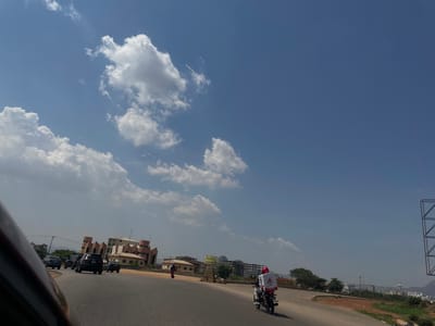 The image depicts a bright daytime street scene, likely in Crescent, Nigeria, under a clear blue sky dotted with fluffy white cumulus clouds. The perspective appears to be from a moving vehicle.
In the mid-ground, a person rides a black motorcycle traveling on a paved road. The motorcycle is equipped with a white delivery box on the rear, which has red lettering clearly displaying "Mr. ECO". The rider wears a red helmet. To the right of the motorcycle, a person in a long, reddish garment walks along the side of the road.
The road curves gently, with various buildings lining the horizon. On the right side of the frame, several black cars, including SUVs and sedans, are visible on the road, some appearing to be parked. Prominent among the buildings is a multi-story structure featuring a distinctive architecture with reddish-brown and white facades, characterized by triangular or jagged rooflines and multiple column-like structures. In the distant background, a cityscape with numerous other buildings is discernible.
To the far left, a large, empty metal frame, possibly for a billboard, stands tall. Green trees and patches of vegetation are scattered along the road and around the buildings. A notable detail in the background is a yellow banner or sign displaying the text "<<<DEPRESSION>>>" in large, dark letters, mounted on a structure further down the road. The overall scene suggests a busy urban or semi-urban environment with ongoing activity.