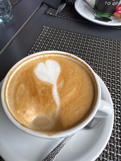 This is a close-up, overhead shot of a white cup of latte coffee with latte art on a white saucer. The coffee has a creamy froth with a light brown swirl pattern, resembling a leaf or heart. The cup is sitting on a dark woven placemat, which is on a dark table. To the left of the coffee cup, on the same placemat, is a small white saucer containing a black cylindrical object with green lettering and a silver spoon. Visible text on the black object reads "TO GO". Part of a silver spoon is also visible on the saucer to the right of the coffee cup. The lighting suggests it is daytime, possibly outdoors or in a well-lit indoor space. The setting appears to be a cafe or restaurant, as indicated by the tableware and placemat. No people are visible in the image. The overall mood is relaxed and suggests a moment of enjoyment. The location context provided indicates this scene is in La Vila Joiosa, Spain.