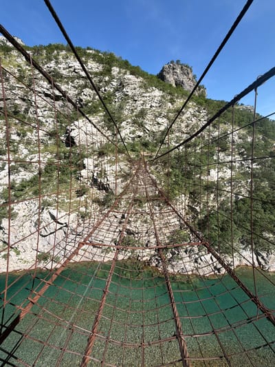 A vertical, eye-level shot captures a rustic, narrow suspension bridge constructed from heavily rusted steel cables and a wire mesh grid, extending across a deep river gorge. The perspective looks directly along the bridge, which tapers sharply into the distance, accentuating its length and the significant drop below. Beneath the bridge, a clear, vibrant turquoise-green river flows over a visible bed of light-colored pebbles and stones. Steep, rugged limestone cliffs, partially covered with sparse green vegetation and trees, rise on both sides of the gorge. A prominent, tree-topped rock formation is visible against the clear, deep blue sky on the upper right. Direct sunlight casts distinct shadows from the bridge structure onto the water. The scene is set in an unknown location in Montenegro.