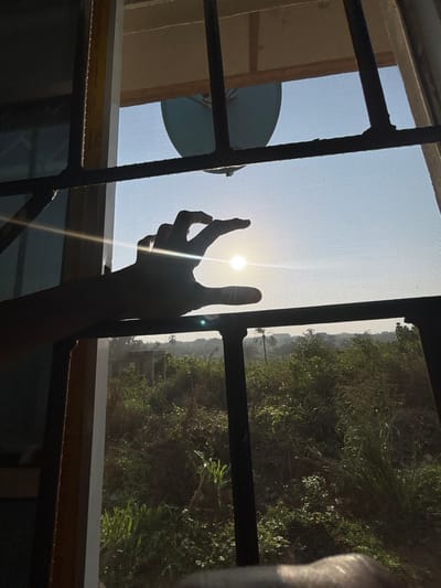 The image shows an outdoor view through a window in the city of Debasco, Nigeria. The foreground displays a dark security grate with horizontal and vertical bars, overlaying an insect screen. A human hand, silhouetted against the bright light, is positioned in the lower left-center, with its thumb and forefinger appearing to encircle a prominent, low-lying sun. The sun emits a strong glare and lens flare. Above the hand, a light-colored, possibly blue or grey, satellite dish is partially visible in the upper-center. The midground and background feature a clear, light blue sky above a dense, green landscape composed of various vegetation, including grasses, bushes, and trees. In the distance, indistinct building structures and additional tall trees are visible. The scene is strongly backlit by the sun, casting most immediate elements into silhouette.