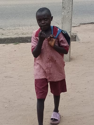 A young, dark-skinned child stands on a sandy ground in Biliri, Nigeria. The child is wearing a pink and maroon striped collared shirt, maroon shorts, and a single pink sandal with the word “Balenciaga” printed on it. A blue and black backpack is slung over their shoulder. The child is looking directly at the camera with a finger near their mouth. A paved road and a concrete curb are visible in the background, along with a concrete pole.