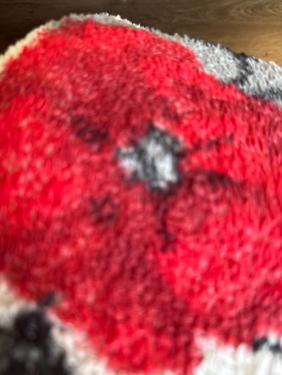 The image shows a close-up, blurred view of a fluffy, red, white, and black patterned textile, possibly a rug or blanket. The pattern appears to be abstract or depict a stylized image, with large red areas and interspersed white and dark grey/black patches. The texture is clearly visible due to the softness and fuzziness of the material. There are no people or specific identifiable objects present in the immediate foreground. The background is dark and out of focus, with a hint of wood grain suggesting a floor or furniture. The location is the city of Ridabu, Norway, but no specific landmarks or environmental details relevant to that location are discernible from the image itself.