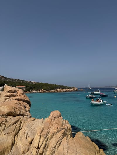 The image depicts a clear blue sky above a picturesque scene in Punta Sardegna, Italy. In the foreground, large, rugged, light-brown rocks dominate the left side of the frame, with textured surfaces and deep crevices. Beyond the rocks, a vibrant turquoise sea stretches out, its clarity allowing the seabed to be partially visible. Scattered across the water are several boats of various sizes, including inflatable dinghies with people in them and larger yachts. Some of the people are casually dressed, suggesting a relaxed summer atmosphere. In the middle ground, a small, red-roofed building is nestled among lush green trees and rocky terrain lining the shore. Further in the distance, other boats are visible, and the coastline curves into the background, dotted with vegetation. The overall impression is one of a sunny day in a beautiful coastal location, likely a popular spot for leisure and water activities.