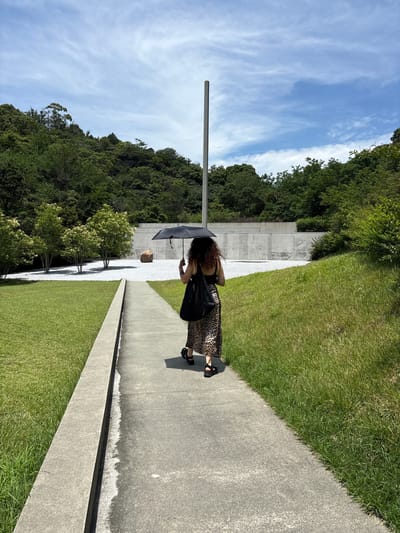 An individual walks along a paved path under a black umbrella. The person is facing away from the camera, wearing a leopard print skirt and dark sandals. They are carrying a black tote bag over their left shoulder. The path is bordered by a manicured lawn on the left and a grassy embankment on the right. In the background, a minimalist concrete structure with a tall, thin pole rises against a blue sky with wispy clouds. The area is surrounded by lush green trees and hills, suggesting an outdoor art installation or park setting. This is the outdoor space at the Chichu Art Museum on Naoshima Island, Japan.