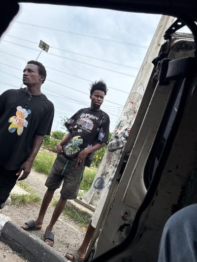 The image shows three young men standing near a curb in Ojomu, Nigeria. One man wears a black T-shirt with a colorful graphic, another a patterned black shirt and gray shorts. The third, partially visible, wears a checkered shirt and light shorts. They are standing next to a vehicle. Overhead are power lines and a solar panel street light. The photo is taken from inside the vehicle.
