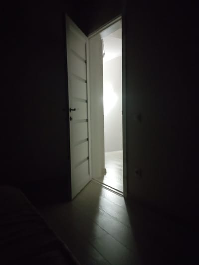 The image depicts a dimly lit interior scene, focusing on an open white door. The door is slightly ajar, revealing a brightly lit hallway or room beyond, casting a strong rectangular shadow on the wooden floor. The door itself has a modern design with horizontal indentations and a silver door handle. To the left of the door, a portion of a dark wall is visible, with a subtle suggestion of a light-colored object protruding from it, possibly a switch or outlet. To the right of the open door, the wall is predominantly dark, with two light-colored square objects visible, likely light switches or electrical outlets. In the foreground, the left side of the image shows a dark, textured surface, possibly a bed or sofa, partially obscuring the wooden floor. The floor is composed of dark wood planks with a visible grain pattern, reflecting the light from the open doorway. The overall atmosphere is one of shadow and contrast, with the bright interior beckoning from the darkness. The specific location in Ialoveni, Moldova cannot be determined from the visual information provided.