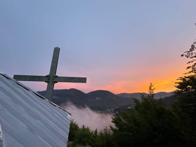 This media file depicts a scenic mountain landscape at dusk in Strazha, Bulgaria. The primary subjects are a large, dark green cross situated in the foreground, to the left of the frame, and the rolling, forested mountains that form the backdrop. A corrugated metal roof extends from the bottom left corner, partially obscuring the base of the cross.
The sky displays a gradient of colors, transitioning from a pale blue at the top to soft pinks and oranges near the horizon, indicating sunset or twilight. A thin layer of fog or mist blankets the valley below the mountains, obscuring the details of a small town or village nestled within. The dense green foliage of trees frames the lower right portion of the image, adding depth and texture to the scene.
The overall mood is serene and contemplative, with the dramatic colors of the sky and the imposing presence of the cross suggesting a moment of peaceful reflection or a spiritual gathering. The time of day is clearly evening, and the weather appears clear with some atmospheric moisture in the valley. There is no visible text in the image.