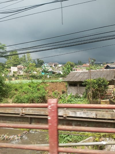 A landscape view with a foreground of a rusty bridge railing above a polluted creek. Beyond, a small village nestled in greenery, with houses having tiled roofs and various vegetation. Dark, ominous clouds fill the sky, suggesting an approaching storm. Utility lines are visible across the top portion of the image.