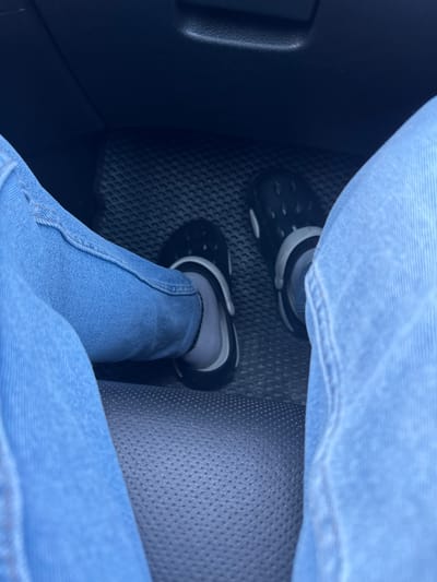 The image shows a close-up view of a person's legs and feet inside a vehicle. The person is wearing blue jeans and black crocs with grey socks. The feet are positioned on the car's floor mat, which appears to be black and textured. The lighting suggests it is daytime. The background shows part of the car's interior, including a dark dashboard and seat. This image was taken in the city of Trușeni, Moldova.
