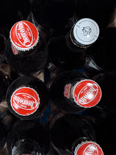 The image shows a close-up view of several glass bottles filled with dark liquid, likely soda, and arranged closely together. The bottles have different colored caps. Three bottles have bright red caps with white lettering that reads "Cheerwine" and "Since 1917," while one cap is white and shows the inscription "Est. 1891" and "Twist Off". The bottles are densely packed, obscuring their contents. The image is taken from an above, angled perspective. The location of the image is unknown and not inferable from the provided data.