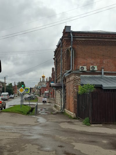 The image depicts a street scene in Kungur, Russia, under an overcast sky. A two-lane road runs through the center of the frame, with cars parked along the right side. A brick building with an ornate facade and a metal roof dominates the right side of the image. There are two outdoor air conditioning units mounted on the brick wall. A dark metal gate is visible in front of the building.
On the left side of the road, there is a sidewalk with pedestrians walking. A traffic light shows a green signal. Several road signs are present, including a pedestrian crossing sign and a diamond-shaped warning sign. A van and other vehicles are on the road. In the background, a golden-domed church stands against the cloudy sky. The wet pavement suggests recent rain.