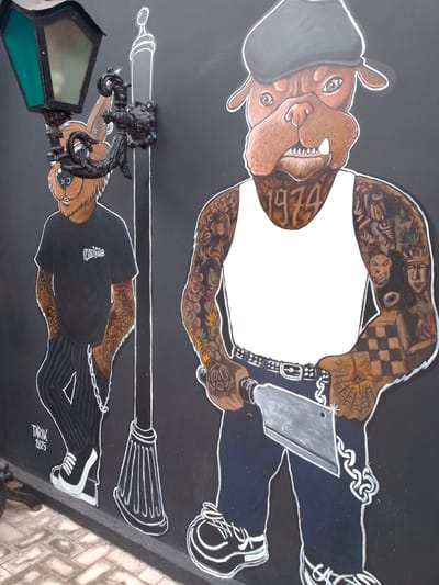 The image is a mural on a black wall, depicting two cartoonish anthropomorphic dogs in a street art style. The mural is located on a wall in Cuiabá, Brazil, likely in an urban setting.

On the left, a dog with a brown, furry face, wearing a black t-shirt with a white logo, dark pinstripe pants, and white sneakers, stands leaning against a lamppost. A silver chain hangs from the belt loops of its pants. The lamppost itself is depicted in black and white, with a black wrought-iron decorative element holding a green-paned lantern. The name "TARIK" and the year "2025" are written in white at the bottom left of this figure.

To the right of the lamppost, a larger dog, possibly a bulldog, is depicted wearing a grey cap, a white tank top, and dark pants. This dog has numerous tattoos covering its arms and neck, including the number "1974" on its throat. It is holding a large, silver cleaver in its right hand. The cleaver has a distinct checkered pattern on the blade. A belt with a buckle is visible around its waist. Both dogs have their outlines in white. The background is a solid black wall.

The ground in front of the mural is a paved area with light-colored stones. The lighting suggests an outdoor environment.