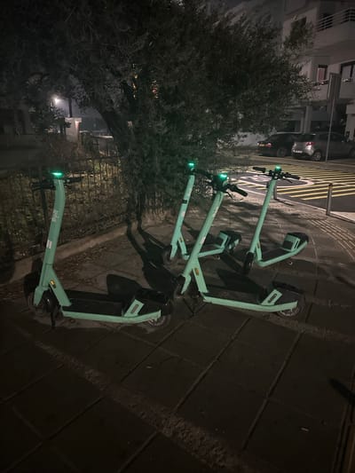 The image depicts an outdoor urban scene at nighttime, possibly a street or sidewalk area. The primary subjects are four light green electric scooters, all branded "Bolt," which are parked in a row along a paved surface. Their front lights and possibly some side indicators are illuminated with a green glow, standing out against the dark environment.

To the left of the scooters, a large, dense tree with dark foliage occupies a significant portion of the frame. Beyond the tree and further down the street, a dark-colored car is parked, and parts of a building with visible windows can be seen. The ground where the scooters are parked appears to be a dark, paved sidewalk, possibly made of large tiles or asphalt, with some reflective surfaces hinting at residual moisture or uneven texture. Shadows cast by the scooters are visible on the ground to their right, indicating a light source coming from the left or above.

In the background, behind the scooters and across what appears to be a street, there are clear yellow and white road markings on a dark asphalt surface, suggesting a designated area or traffic guidance. The overall setting is consistent with a city or town environment, given the presence of scooters for hire, a parked car, and buildings. There is no human activity taking place; the scene is static, focusing on the parked vehicles and their immediate surroundings under artificial lighting. The text "Bolt" is prominently visible on the body of each scooter.