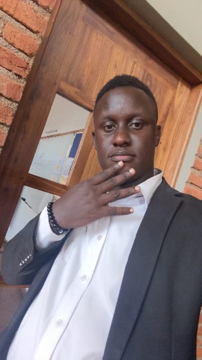 An adult male, with dark skin and short, dark hair, is depicted in a portrait photograph taken indoors in the city of Karatu, Tanzania. He is wearing a white collared button-up shirt, open at the collar, beneath a dark blazer. A black beaded bracelet is present on his left wrist. His left hand is raised to his lower face, with fingers lightly splayed. The subject maintains direct eye contact with the camera, displaying a neutral facial expression. The background features a reddish-brown brick wall on the left side and a dark wooden door frame or paneling centrally and to the right, which incorporates a reflective or transparent section revealing a light-colored, indistinct interior space.