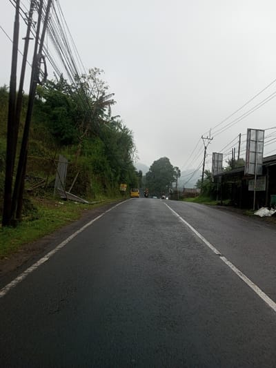 This image displays a two-lane asphalt road within the city of YonPhoogla, Bhutan, under an overcast sky. The road surface appears damp. The perspective is from ground level, looking forward along the road.
To the immediate left, multiple dark utility poles support numerous black electrical and communication cables. Adjacent to the poles is a steep, green embankment densely covered with vegetation, including broad-leafed plants and banana trees. A section of corrugated metal sheeting is propped against the lower part of the embankment. The left edge of the road is defined by a solid white line, bordered by a narrow dirt and grass shoulder.
The asphalt road features a broken white line separating the two lanes. In the mid-distance on the left lane, a yellow van is observed, facing away from the viewer. Behind the van, a person on a dark motorcycle or scooter is also heading away. Further down the road, a second person on a motorcycle or scooter is visible in the right lane, proceeding in the same direction.
On the right side of the road, additional utility poles with overhead lines are present. A series of three rectangular, light-colored signs with dark frames are affixed to structures alongside the road. A dark building is partially visible behind these signs. The right edge of the road is marked by a solid white line.
In the background, a large green tree stands centrally, with other trees and mountainous terrain visible behind it, partially obscured by haze. The sky is uniformly light grey and overcast. No pedestrians are visible.