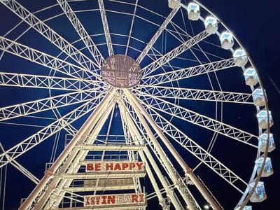 The image shows a large Ferris wheel at night. The Ferris wheel is illuminated with white lights and has many spokes radiating from the center. Several gondolas are visible, each lit with blueish light. In the foreground, there are large red letters on white platforms spelling out "BE HAPPY" and "YOU ARE IN BARI". The background is a dark blue night sky. The Ferris wheel appears to be part of a temporary installation or amusement park, as suggested by the signage. The location is stated to be the city of Porrentruy, Switzerland, however the signage clearly indicates "BARI", which is a city in Italy.